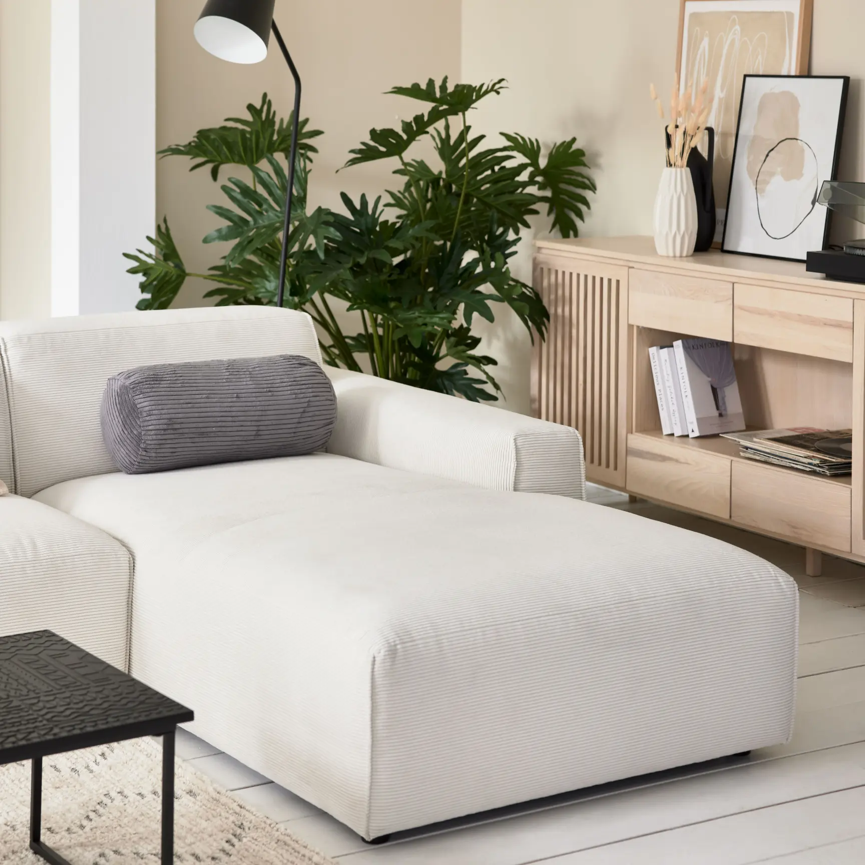 Modernes Wohnzimmer mit beiger Recamiere, grauem Nackenkissen, Sideboard aus hellem Holz und dekorativen Wohnaccessoires – stilvolle Einrichtung im skandinavischen Design mit grüner Zimmerpflanze und Wandkunst.