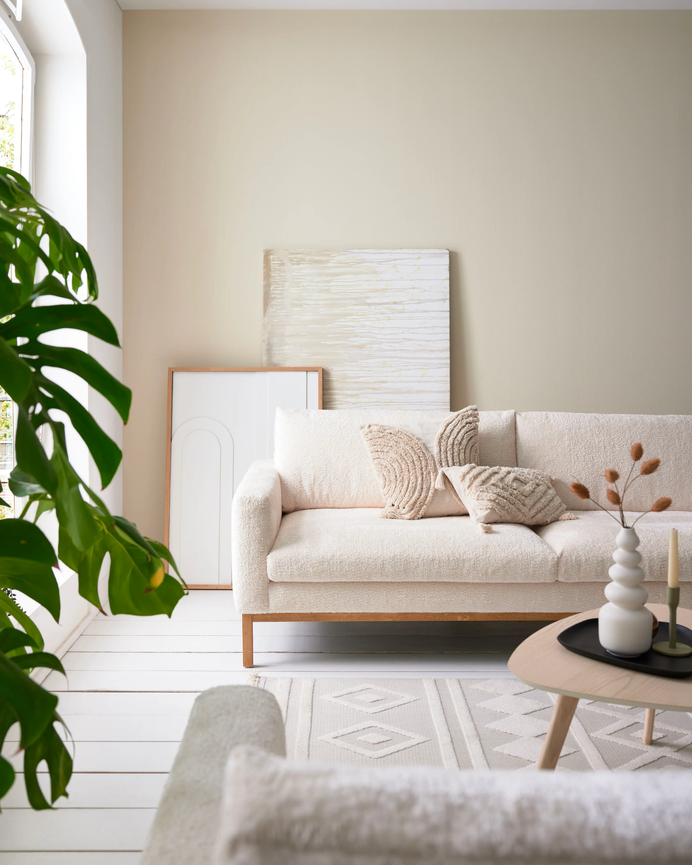 Skandinavisches Wohnzimmer mit beigem Sofa, strukturierten Kissen, runder Vase und großem grünem Blattwerk – natürliche Einrichtungsidee mit harmonischer Farbgestaltung.
