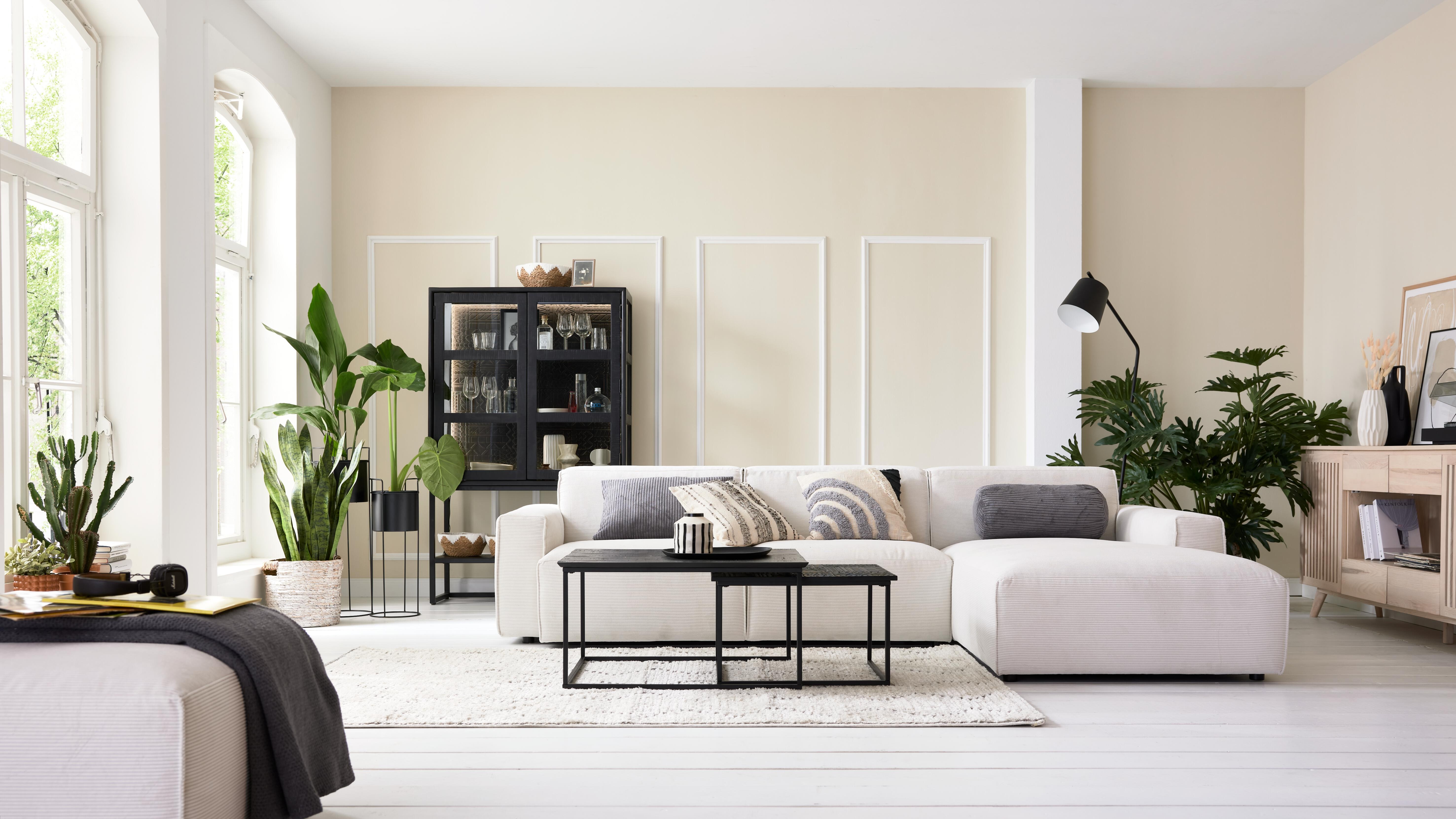 Wohnzimmer mit cremefarbenem Cord-Ecksofa, schwarzen Couchtischen und Pflanzen vor beigefarbener Wand