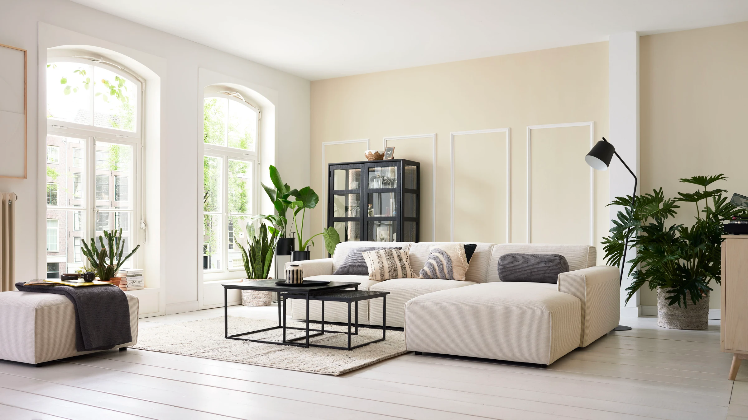 Helles, modernes Wohnzimmer mit großem Roomio Ecksofa in Beige, schwarzen Couchtischen, Glastürenschrank im Industrial-Look und dekorativen Zimmerpflanzen.