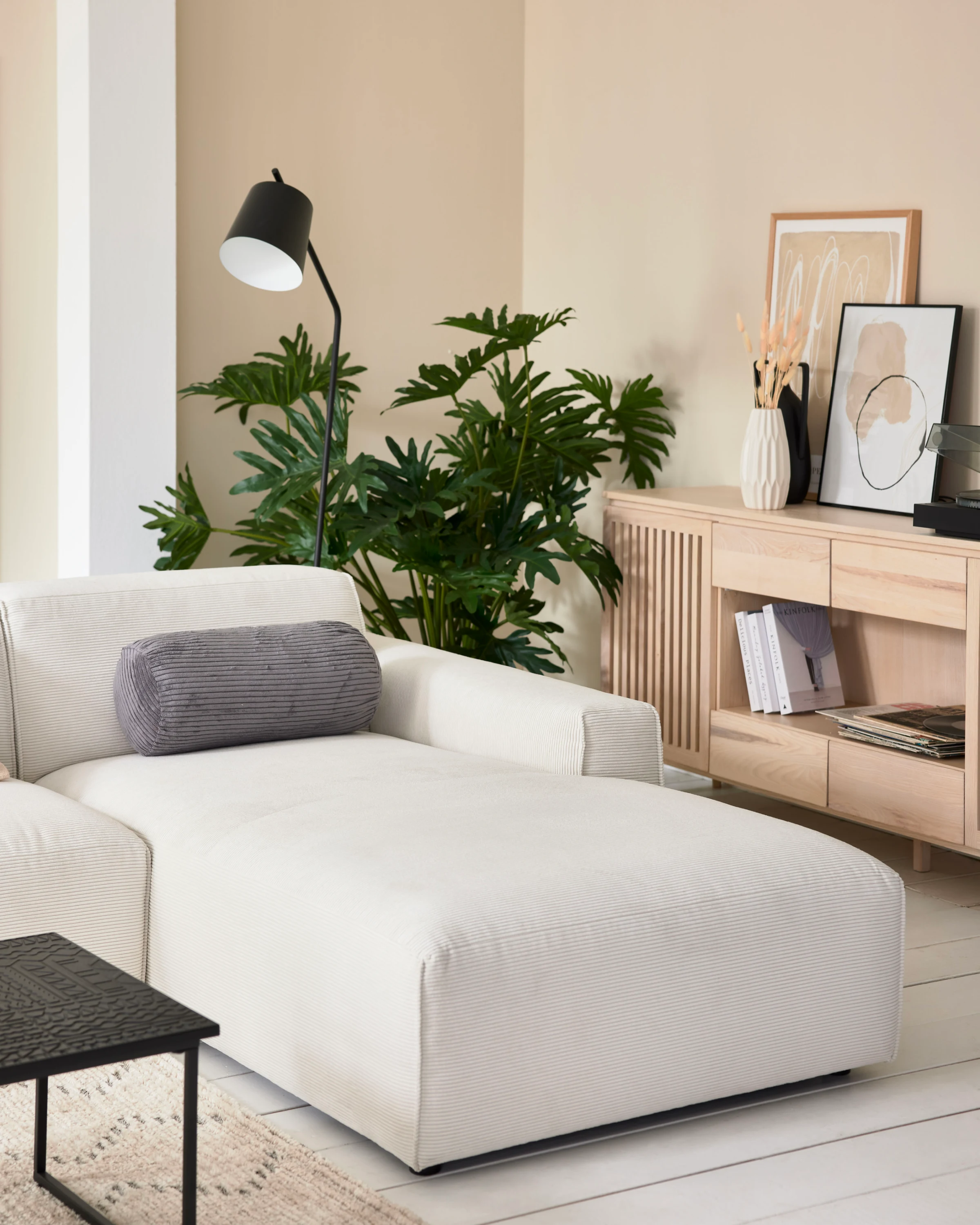 Beigefarbene Recamiere aus der Roomio Serie No. 4100 mit grauem Kissen, Holz-Sideboard, großem Wandbild und grüner Pflanze – elegantes, modernes Wohnzimmerdesign.
